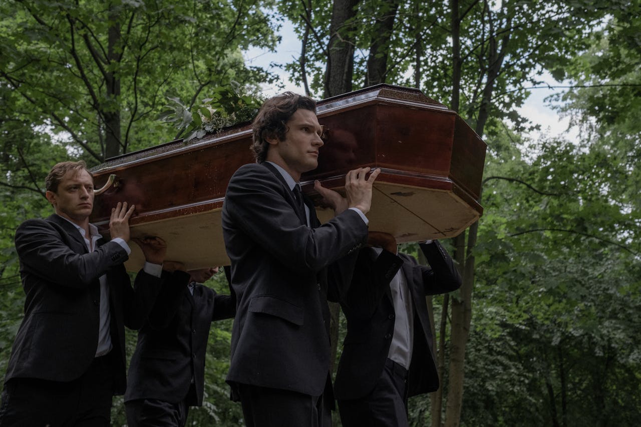 about-02 Men in black suits solemnly carry a coffin through an outdoor forest setting.