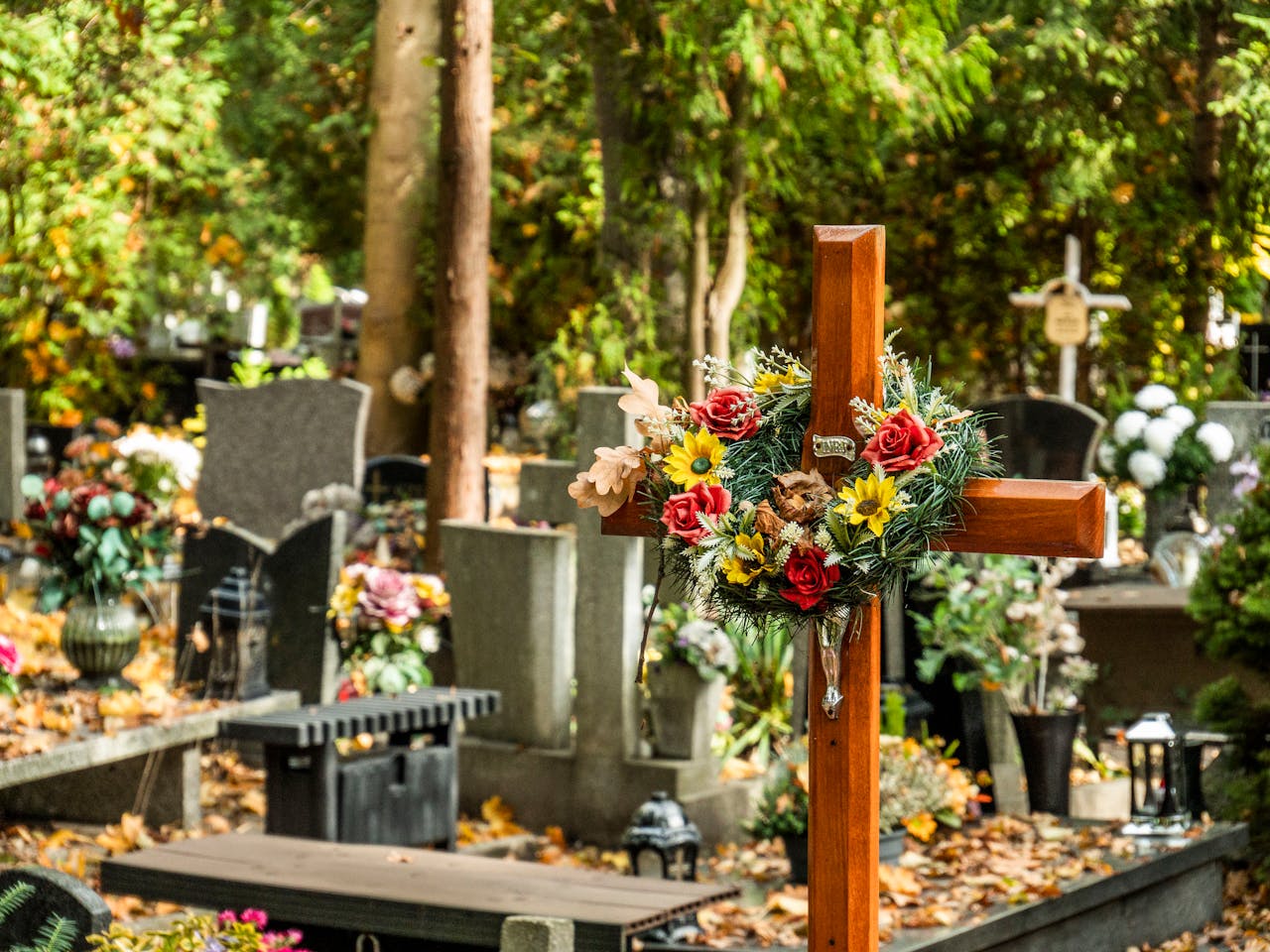 approach-img A peaceful cemetery with a wooden cross adorned with colorful flowers.